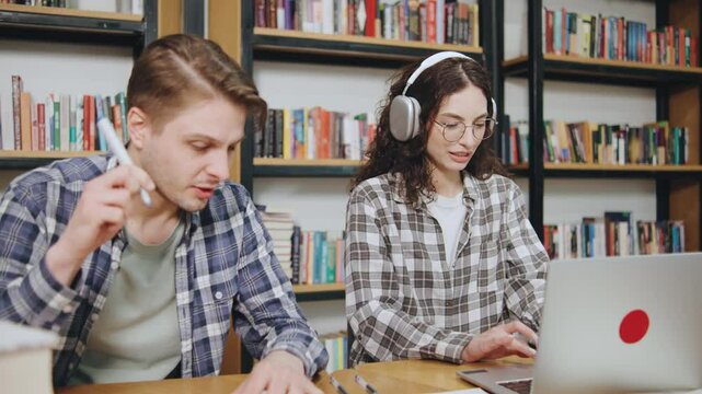 Young adults engaged in study session together at a library surrounded by books in the evening, focused on assignments and technology
