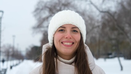 Portrait of woman in warm white knitted hat outdoors in winter. Young woman smiling and enjoying...