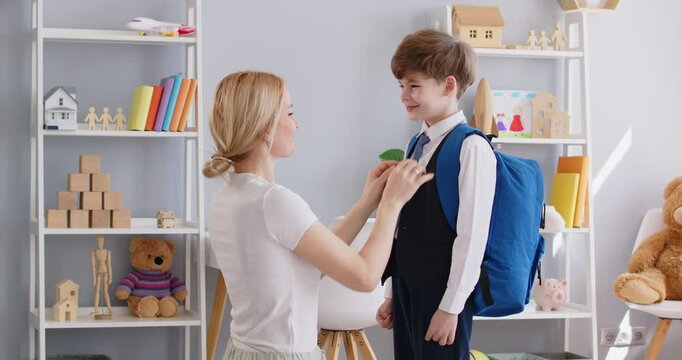 Caring mother carefully straightens her sons clothes before sending him to school. Little boy in childrens room happily tells something while his mother fixes his tie, shirt and backpack. 4K video.