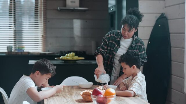 An adult Black woman pouring milk into the bowls of her two school-age sons, who are sitting at the kitchen table and then start to eat cereal with milk in a bright, modern kitchen.