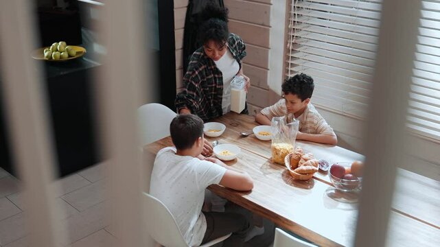 A Latino mother in her 30s approaches a wooden dining table, preparing breakfast. She pours milk into cereal bowls for her two school-aged sons, who sit waiting patiently to eat.