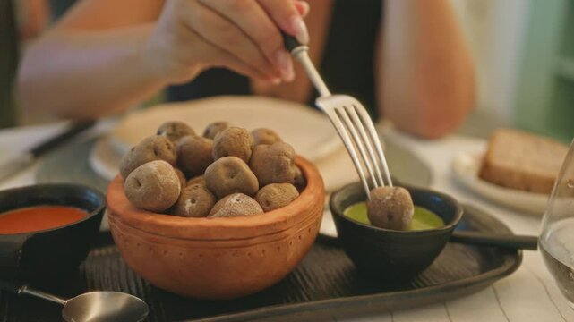 Young woman dining indoors at a fancy restaurant, dipping a potato in green sauce with other dishes on the table