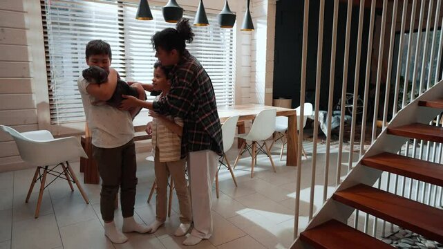 An adult Black woman and her two school-age sons are standing in the kitchen, with one of the boys holding a French Bulldog and then giving it to his mom in a bright and modern living space.