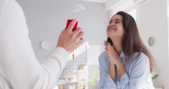 Young surprised happy woman is happy about the marriage proposal from boyfriend. Man holding red gift box with ring proposing to excited girlfriend at home. Couple engagement concept. 4k video. - Powered by Adobe