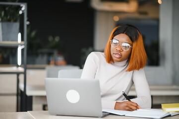 Obraz premium Young African American student girl studying in university, working on research paper, sitting at table with open books
