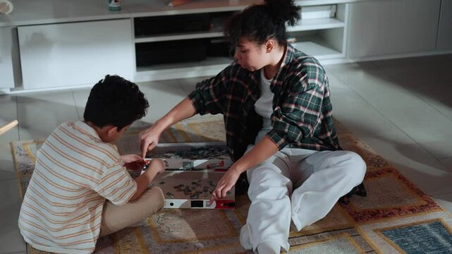 A Latino mother in her 30s and her little son sit together on a patterned rug in a living room. They assemble a jigsaw puzzle, while a TV screen in the background shows a fire.