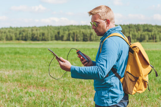 A young man holds a power bank in his hands and charges a digital tablet. Background of sky and field with green grass.