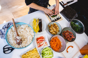 Hands of woman preparing kimbap with bowls and plates of colorful fresh ingredients on white kitchen counter