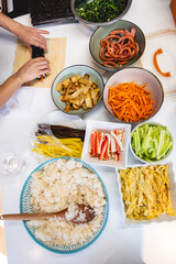 Hands of woman preparing kimbap with bowls and plates of colorful fresh ingredients on white kitchen counter