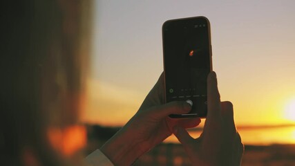 Female traveler photographing dramatic sunset, touch smartphone screen, capturing golden light sweeping across seascape with vibrant coastal horizon. Europe travel.Travel, blogging, weekend, relax  - Powered by Adobe