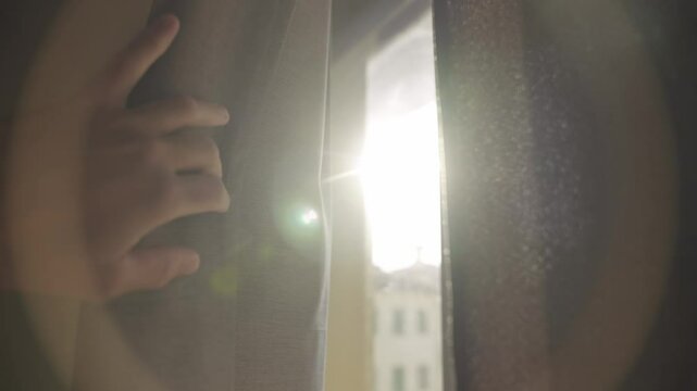 Hand pulls open window curtain to in dark room revealing sunlight shining in. Daylight from outside apartment or home shining bright creating lens flare. Hope and renewable energy or green solutions