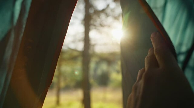 Hand pulls open tent door or fabric to reveal sunlight outdoors in camp site. Camping outside in forest or natural reserve. Close up on man waking up opening tent in early morning to see sun shining
