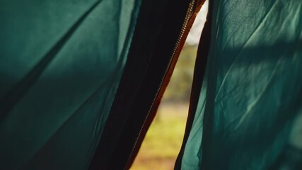 Close up on hand zipping open camping tent to reveal sun shining outside. Early morning in campsite in woods or forest. Person unzips fabric entrance. Warm light creates lens flare. Hand closes hatch