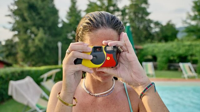 Young woman taking pictures with disposable stills camera in pool party outdoors. Portrait of happy cheerful female in fun party smiling takes film photo towards camera in garden with sun umbrellas