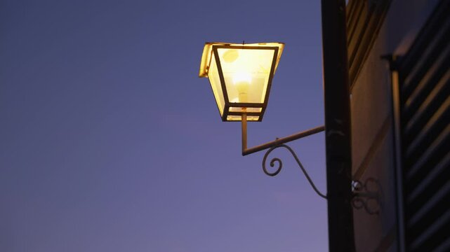Old street lamp lighting and turning on to illuminate dark night at blue hour. Lantern turns on when darkness falls. Blue dark sky. Scary and mysterious. Warm light glowing outside big luxurious house