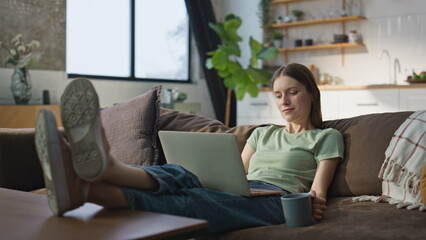 Relaxed woman browsing laptop on knees lying cozy couch with coffee cup home