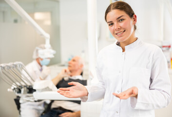 Fototapeta premium Young woman doctor is standing in dental office, clinic employee in white suit uniform near dentists workplace. Doctor enjoys working in profession.