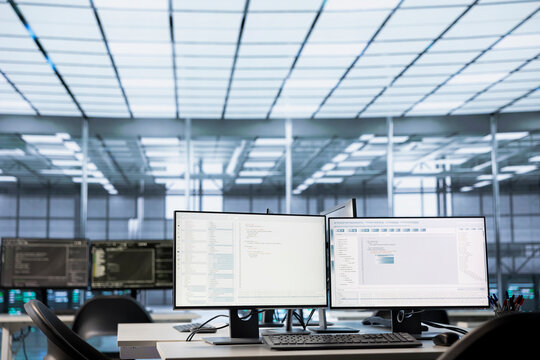 Code running on computer displays in empty data center workspace, used for troubleshooting rackmounts. Binary script running on desktop PC monitors upgrading mainframes systems in server room