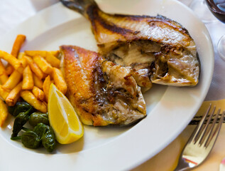 Fried cut dorada fish served on white plate with vegetable garnish and lemon..