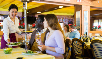 Polite Asian waitress serving ordered dishes to adult biracial couple having romantic date in cozy restaurant..
