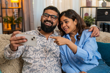 Indian couple man and woman sitting on home sofa, laughing while watching funny social media videos...