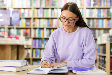 Intelligent female student wearing glasses, sitting at table in library, preparing for lecture or exam, writing notes in notebook, free space