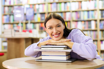 Positive European female student sitting at desk and lying on stack of books in library or book store, looking and smiling at camera, free space