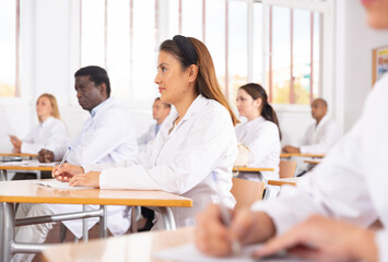 Positive latin american girl in white coat listening to lecture and taking notes in classroom...