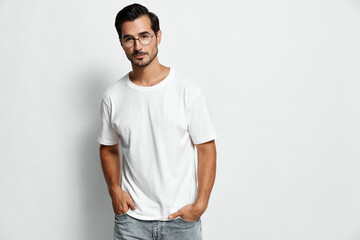 Stylish young man wearing a white t-shirt and glasses stands confidently with hands in pockets against a plain gray background. Fashion lifestyle concept.