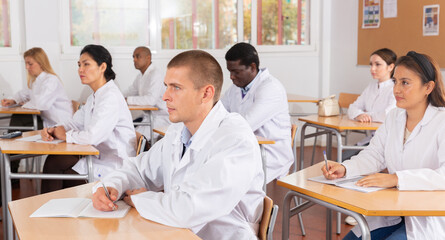 Fototapeta premium Portrait of young man in white medical coat sitting at desk in classroom attending seminar