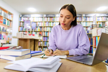 Concentrated female student studying with books and laptop in library, preparing for evaluation...