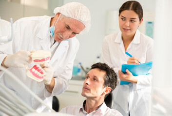 Obraz premium During appointment, mature man dentist and girl nurse communicates with male patient, shows model of jaws and explains mechanism of defect formation.