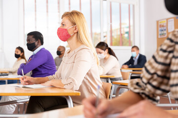 Group of students in protective mask listening attentively to teacher explaining material in classroom