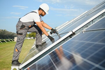 Portrait of Young indian man technician wearing white hard hat near solar panels against blue sky. Industrial worker solar system installation, renewable green energy generation concept