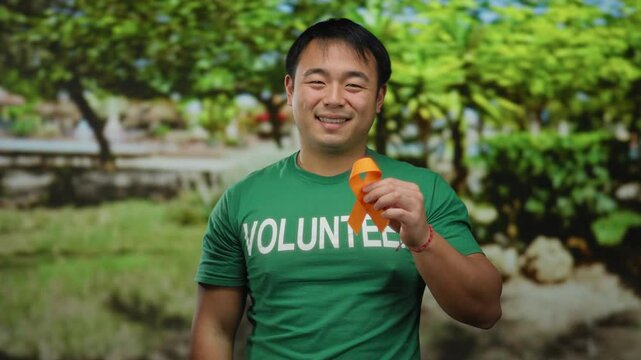 Young man wearing a green volunteer shirt holds an orange ribbon while standing outdoors in a park setting with lush green trees in the background.