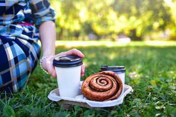 picnic food and drinks on the grass in a sunny park.two coffee cups and a large pastry, with a hand...