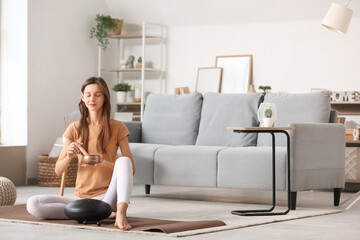 Young woman with Tibetan singing bowl meditating on mat at home