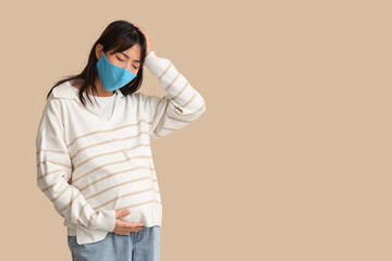 Pregnant Asian woman in medical mask feeling bad on beige background