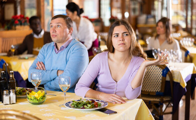 Unhappy adult couple sitting at served table in restaurant frustrated after quarrel during date dinner..