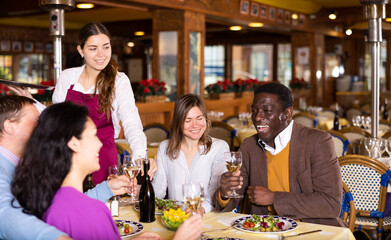 Young waitress serving bottle of wine to cheerful multiracial group of adults having friendly get-together in cozy restaurant..