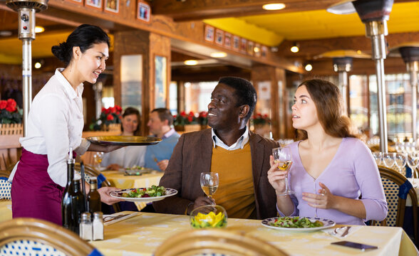 Positive african american man enjoying company of attractive young woman over dinner in restaurant, sitting at table with glass of wine while Asian waitress serving ordered dishes..