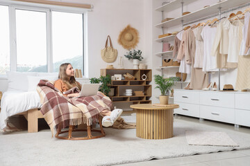 Young woman using laptop and sitting in armchair in bedroom