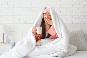 Morning of sleepy young woman with coffee cup under blanket sitting in bedroom