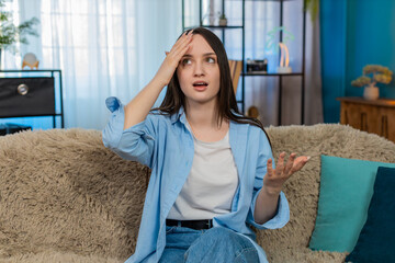 Young woman at home sitting on sofa upset and disappointed doing facepalm gesture, frustrated by...