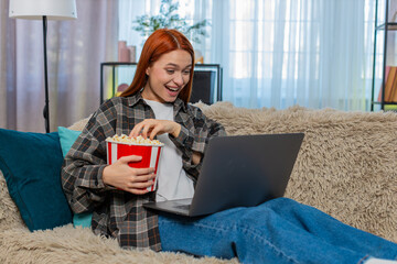 Fototapeta premium Happy woman at home watching movie on laptop and eating popcorn while laughing with joy and excitement. Caucasian redhead girl on sofa enjoying emotional scene, relaxed evening with fun entertainment