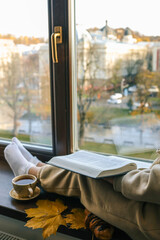 Girl in cozy outfit reading Bible by the window with cup of tea, croissant, and autumn maple leaves, overlooking a sunlit city park in fall