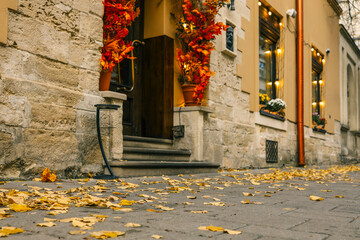Cozy autumn street scene with yellow fallen leaves, stone building facade, warm lights, and red-orange seasonal decorations at the entrance of a cafe or boutique