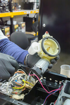 
Technician handling coffee machine gear motor component during internal repair; visible circuit wires and heating system inside technical workspace.

