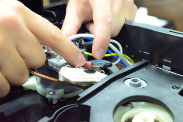 Technician repairing internal parts of a coffee machine, showing close-up of wires, tubes, and components during maintenance or electronic servicing on a workbench.

