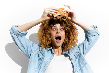 Enthusiastic young woman with curly hair joyfully holding a burger, showcasing excitement for fast...